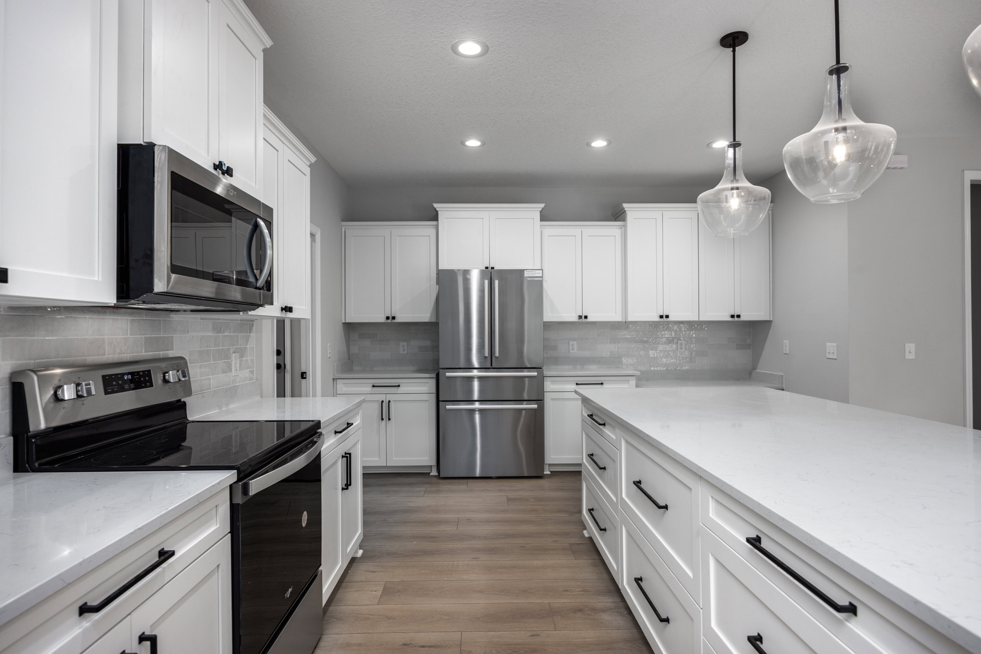 White kitchen with shaker cabinets, stainless steel refrigerator and stove, open microwave door, white countertop with black handles, clear pendant light fixture