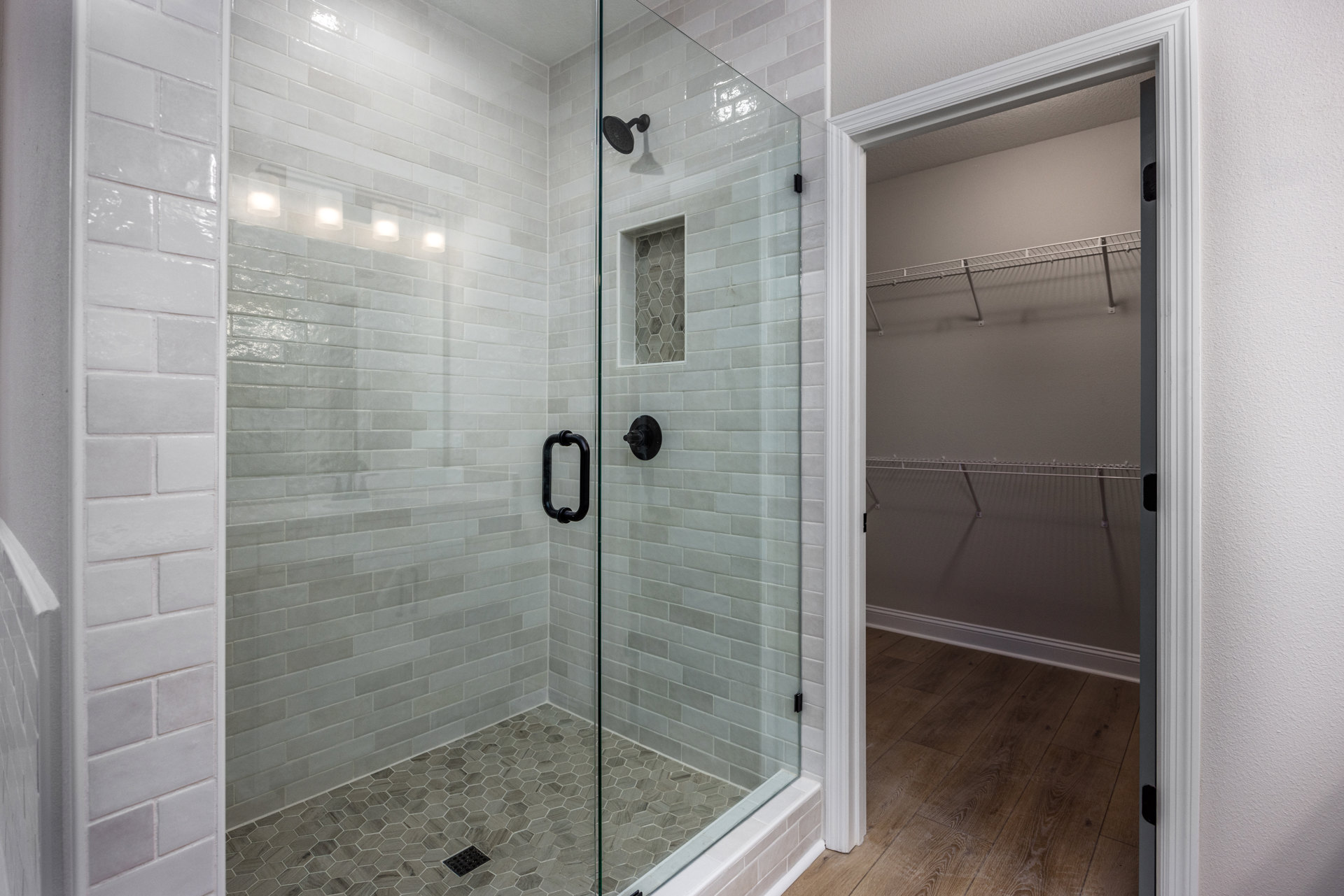 Glass shower enclosure with black hardware, white brick accent wall, tile flooring featuring a black drain grate, and a mounted shower head.