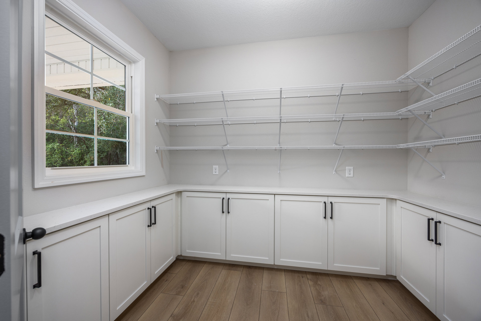 White kitchen with open shelves and cabinets featuring black handles, wood flooring, and a window overlooking green trees.