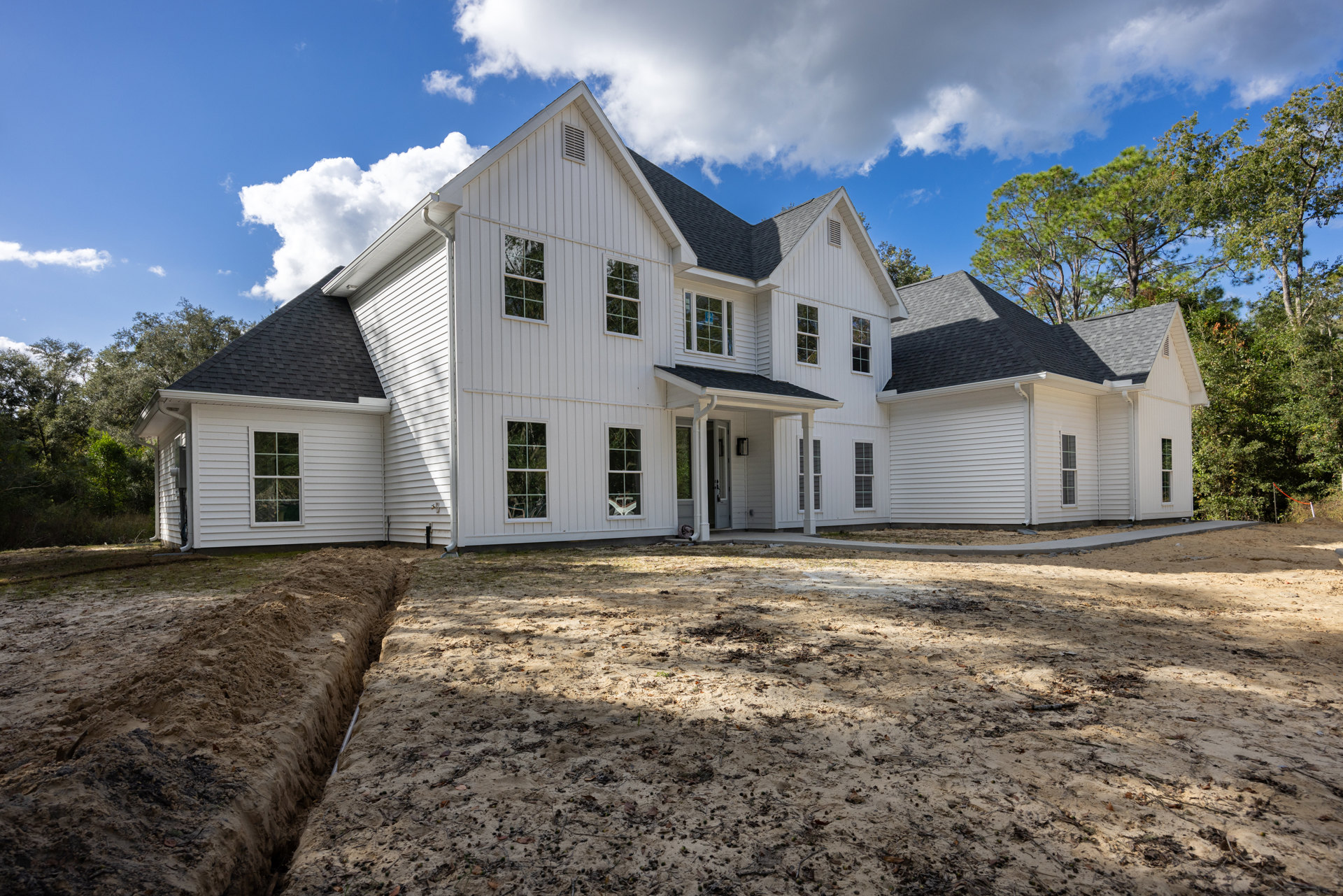 White two-story house with multiple windows, white trim, and a dirt yard scattered with leaves; blue sky with clouds above, trench visible in the foreground, tree reflections in