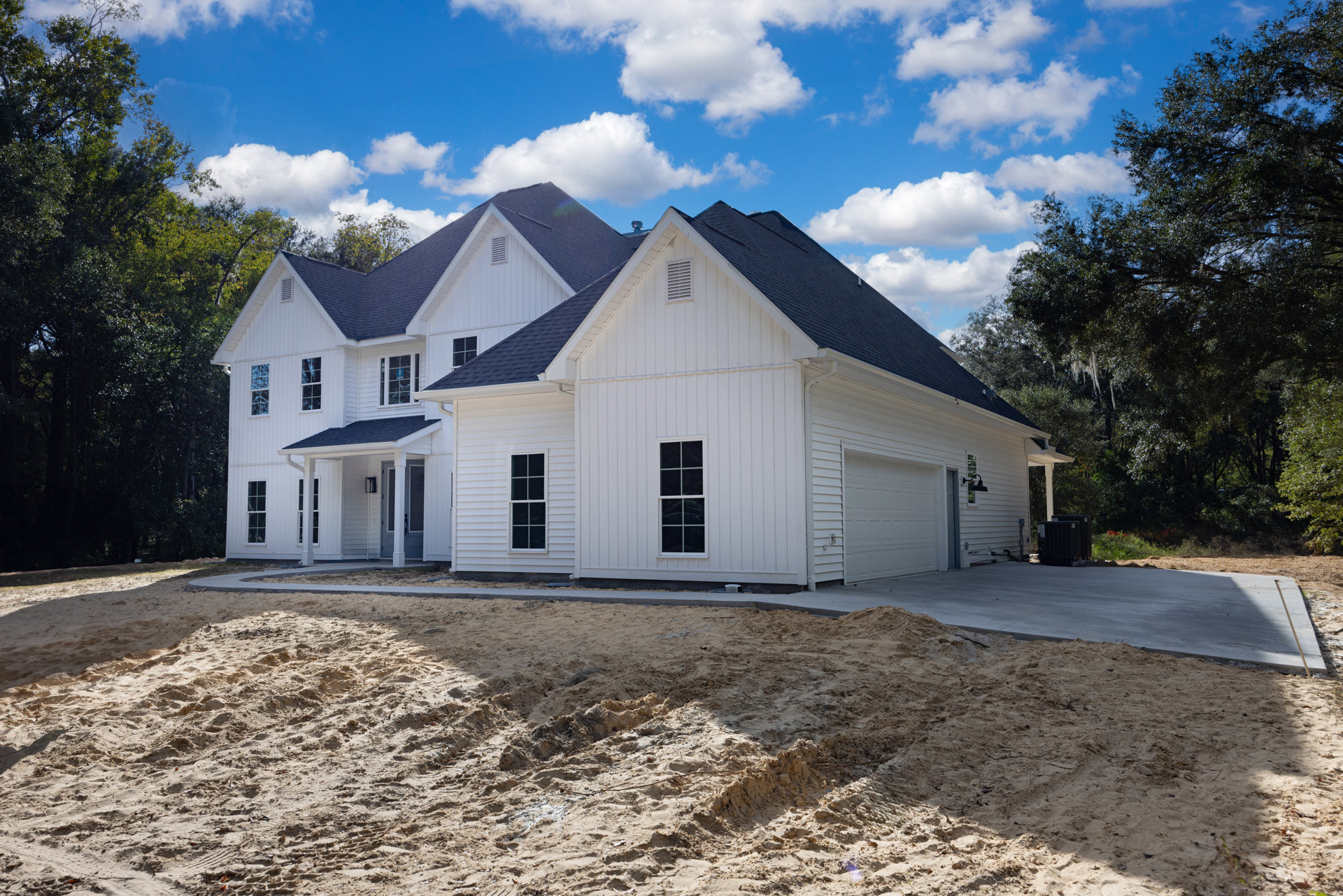 White house under construction with white-framed window, dirt patch in front yard, blue sky and scattered clouds overhead, neighboring houses visible in background