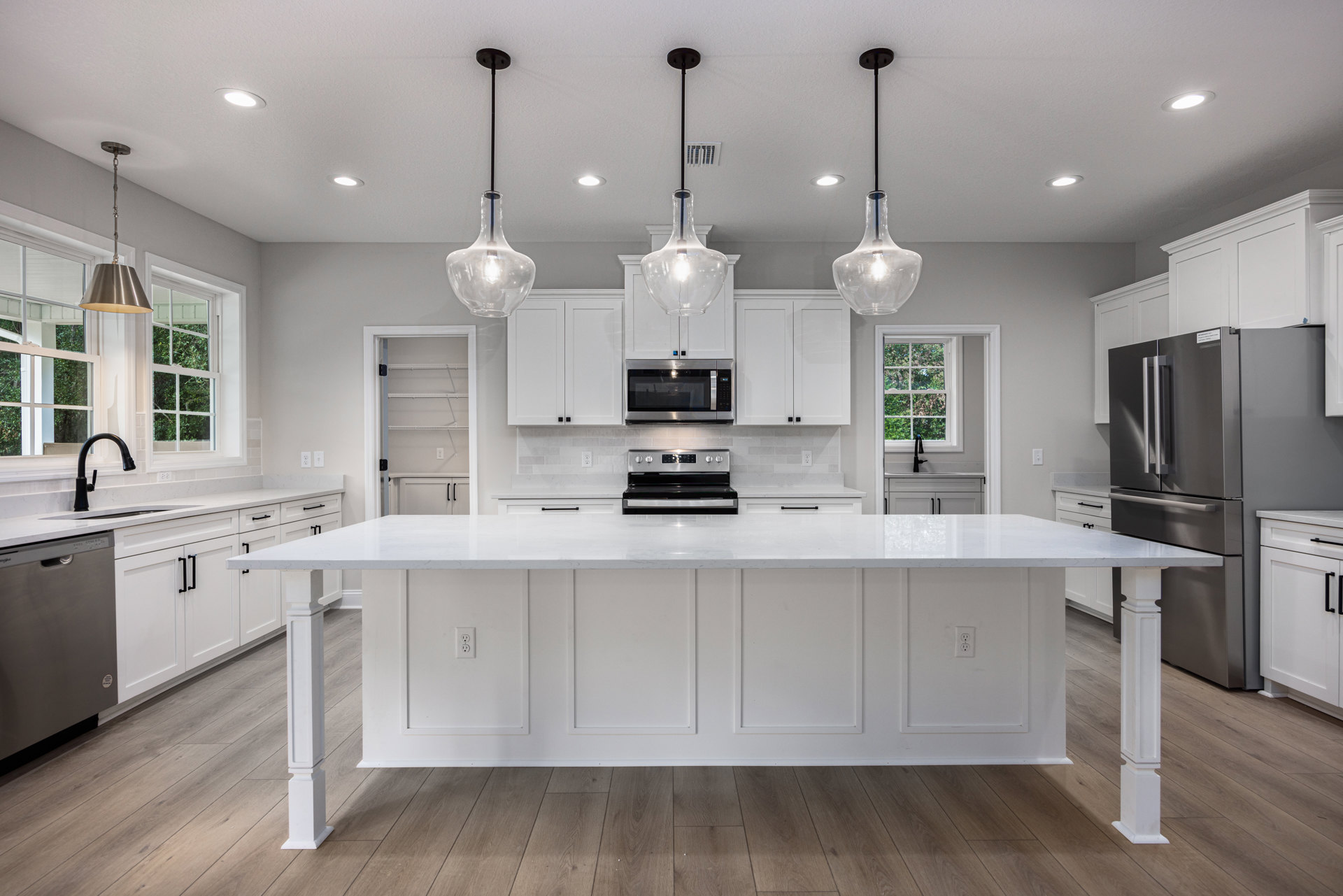 White kitchen with shaker cabinets, quartz countertops, central island, stainless steel microwave and stove, clear glass pendant light, and wood flooring