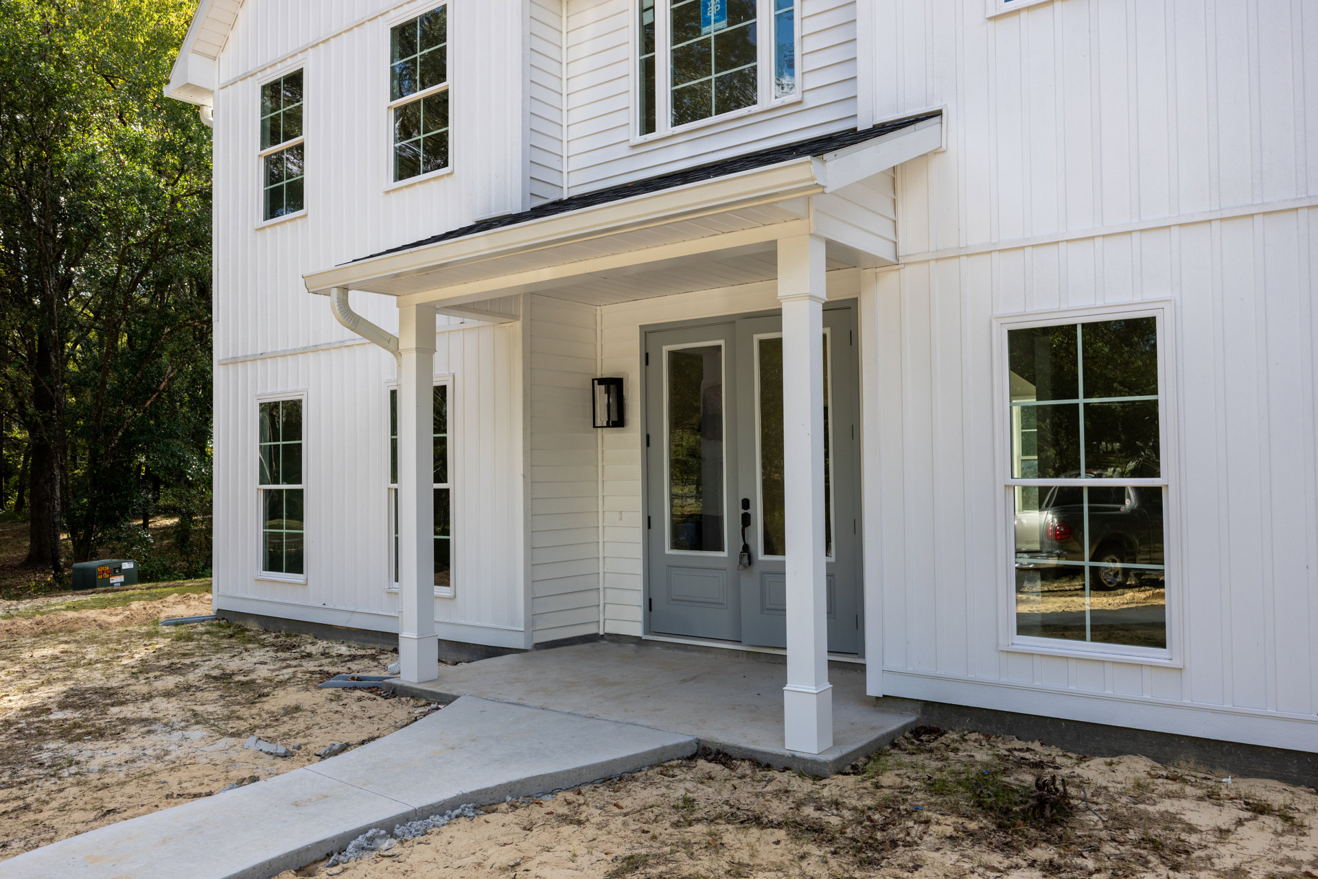 White siding house with two grey doors, white-framed windows, front porch, and green-leaved trees nearby; truck reflected in window.