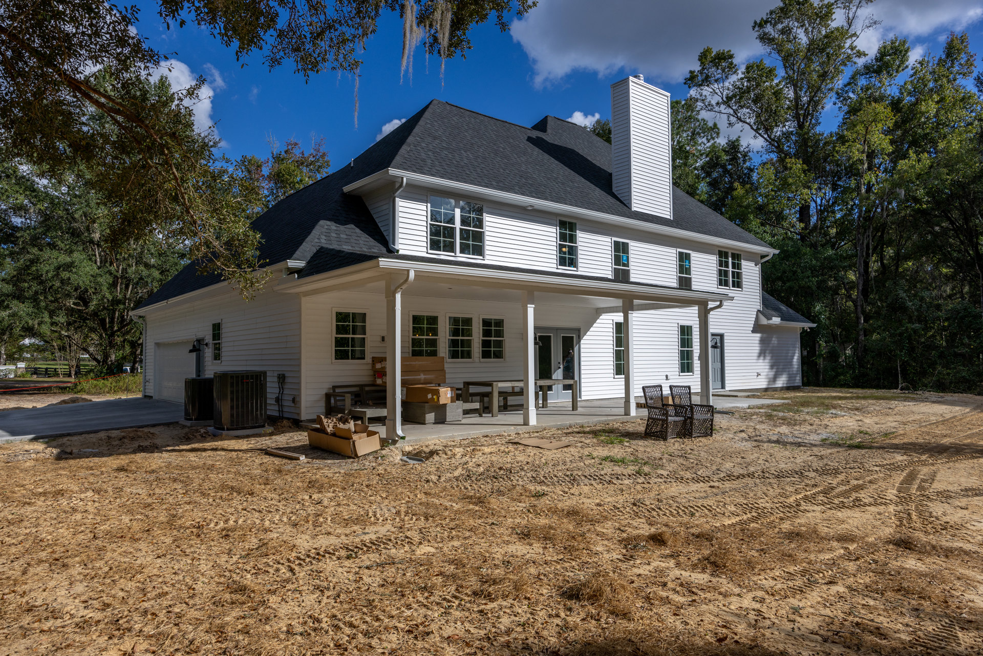 White house with chimney and large yard, wicker chairs on porch, dirt ground with cardboard boxes, white-framed window, trees and plants in background