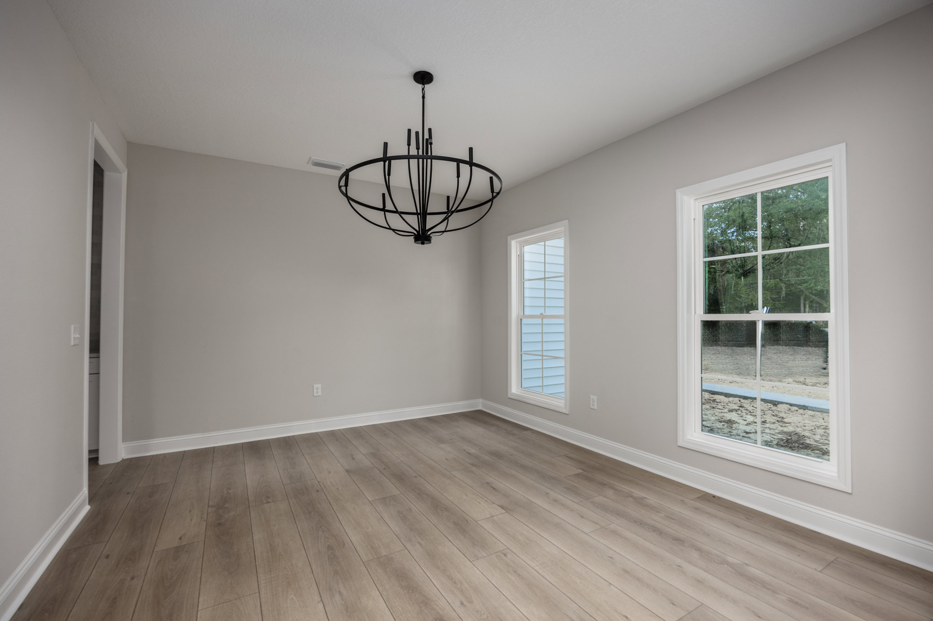 Chandelier with candle-style lights hanging from white ceiling, hardwood floors, large windows framed by white trim, trees visible outside, neutral plaster walls