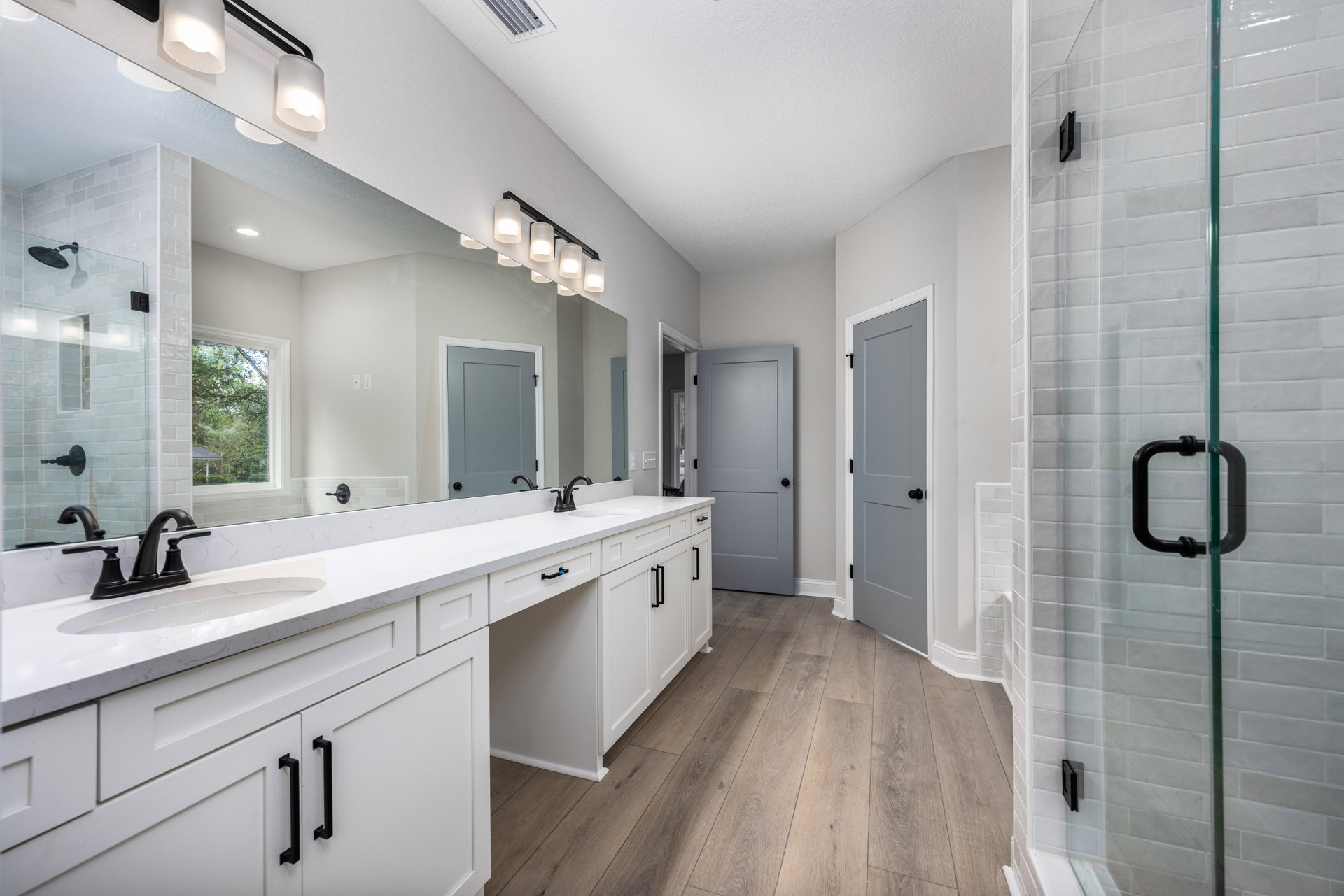 Bathroom featuring a wide vanity mirror, glass shower enclosure with black hardware, white door with black knob, grey cabinetry with black handles, window overlooking trees, light