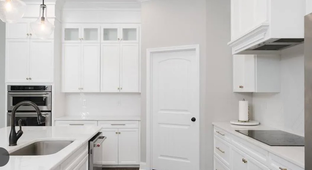 White kitchen featuring shaker-style white cabinets, stainless steel oven with faucet above, quartz countertops, undermount sink, and a door leading to an adjacent room.