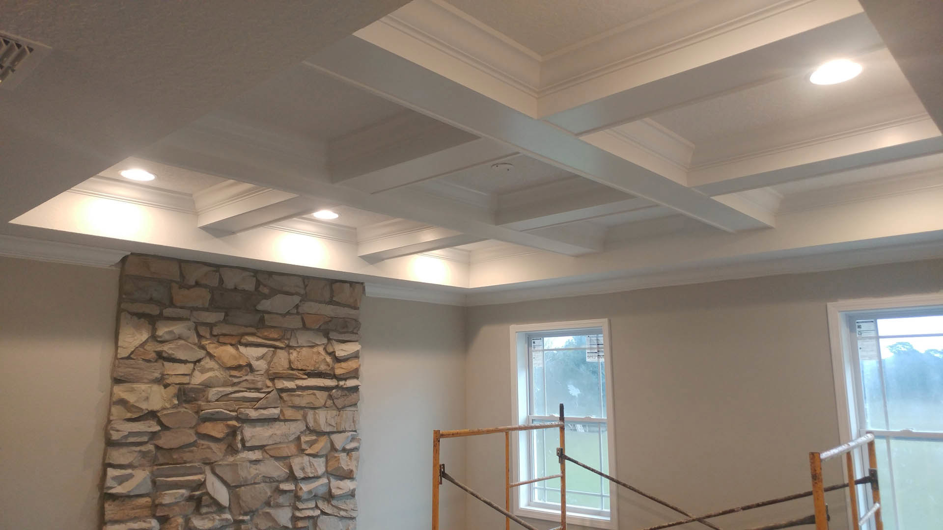 Stone accent wall with white-framed window, white ceiling beams with recessed lights, wood ladder leaning against wall, light-colored floor, window featuring metal railing and