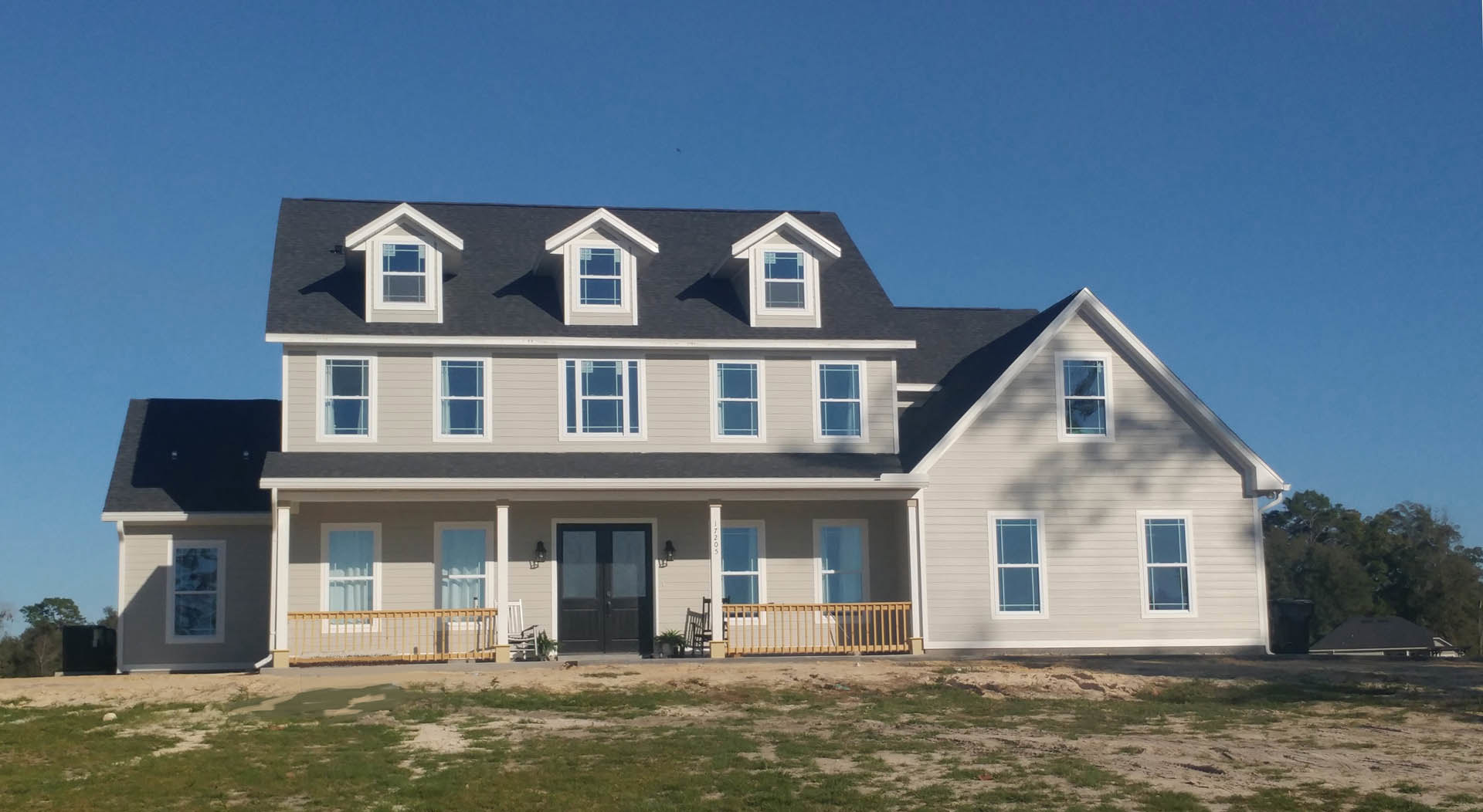 Spacious two-story home with white siding, prominent front porch, double glass entry doors, multiple windows with white frames, manicured green lawn, and clear blue sky overhead