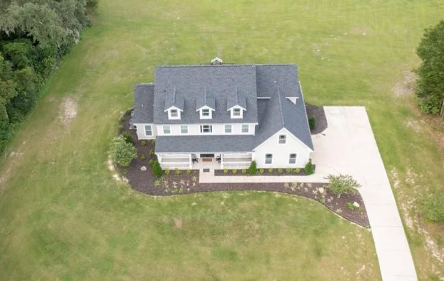 Two-story house with large windows, light-colored siding, front lawn, concrete driveway, and landscaped shrubs and trees