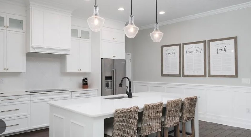 White kitchen island with wicker chairs, white countertops, stainless steel refrigerator, framed pictures on wall, light cabinetry, and modern sink fixtures.