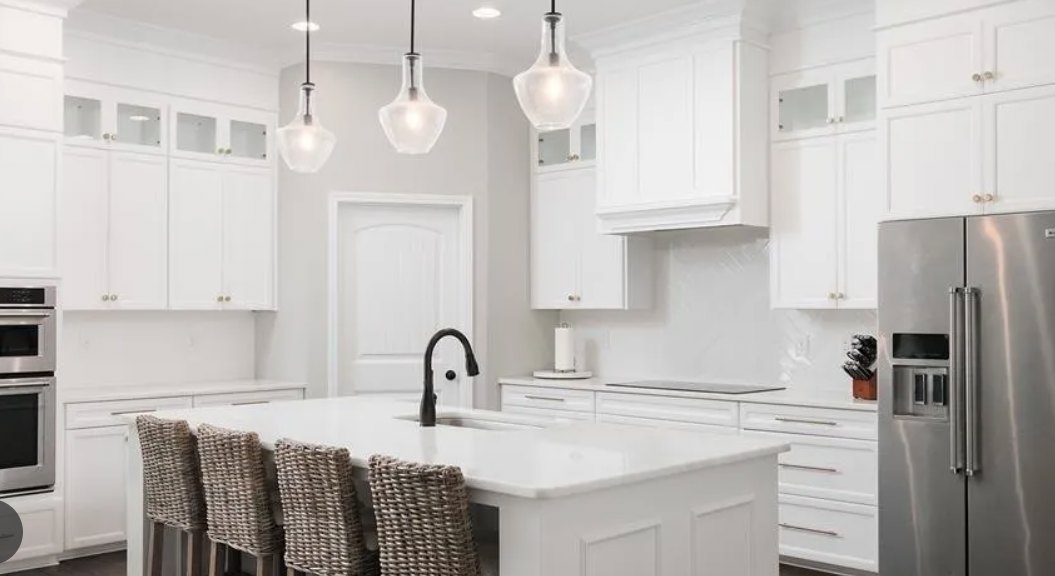 White kitchen featuring a white island with wicker chairs, shaker cabinetry, stainless refrigerator, built-in white oven, and light quartz countertops
