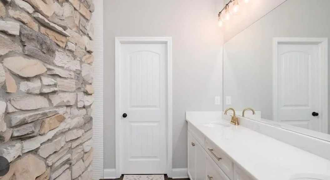 Bathroom with white door featuring black handle, white countertop and gold faucet, stone accent wall, and modern sink.