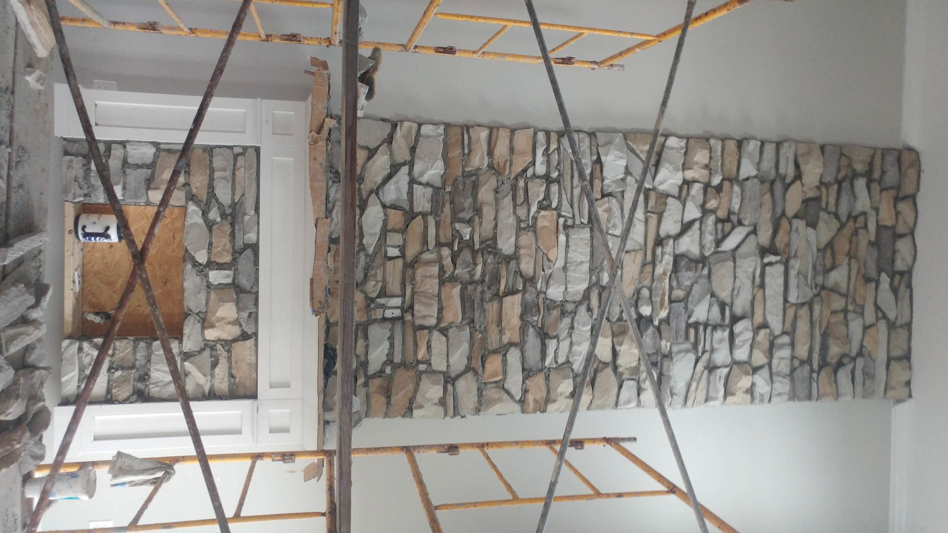 Stone wall under construction with scaffolding, yellow ladder, wooden framing, white can, and various tools and materials scattered indoors.