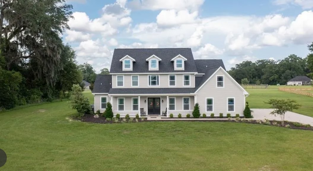 Large two-story house with black roof, expansive green lawn, moss-draped tree, small sapling in dirt patch, and Charles Pinckney National Historic Site visible in the background