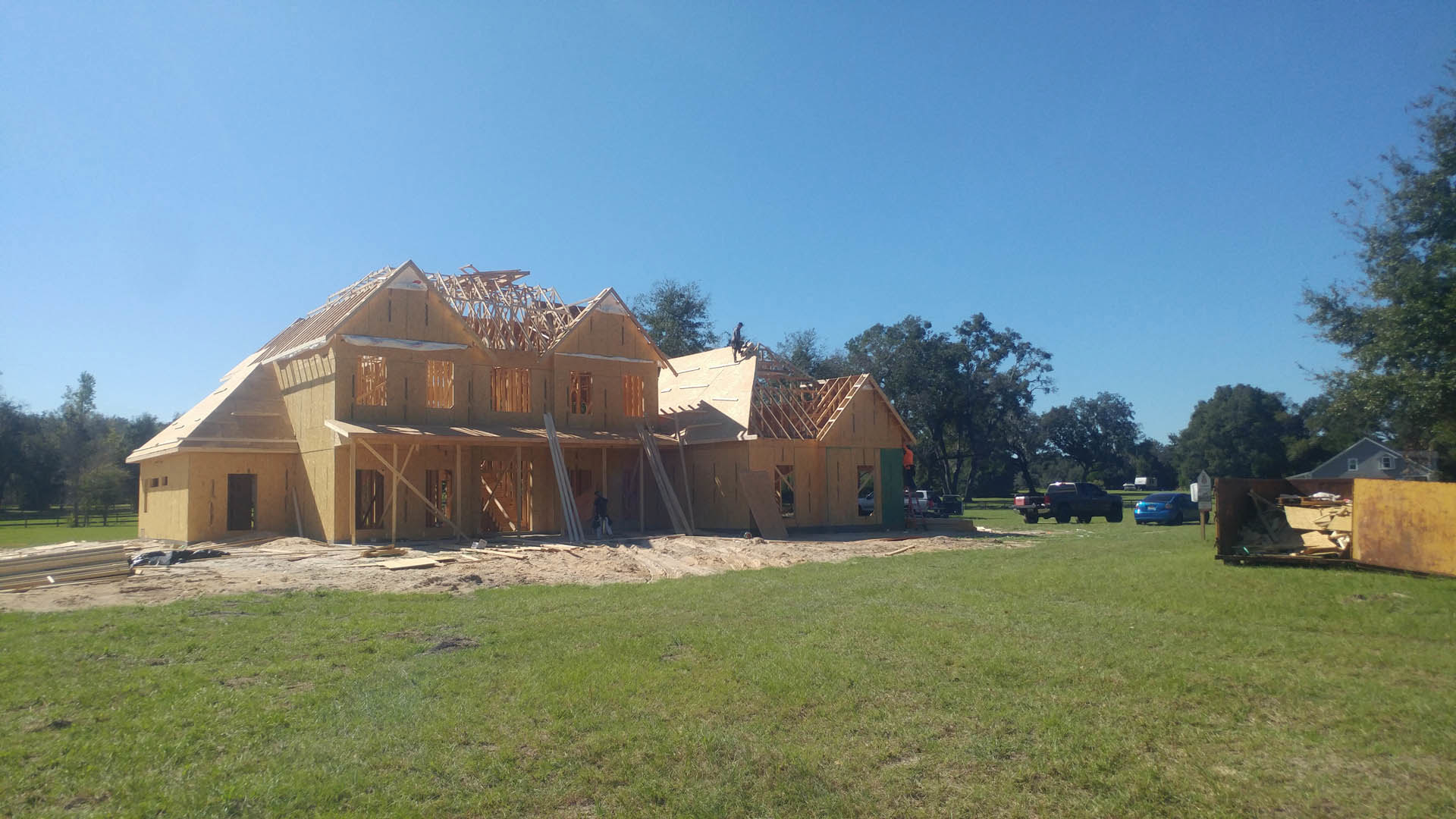 Wood-framed house under construction with exposed roof trusses, surrounded by green grass and mature trees, blue car parked on lawn, worker standing on roof