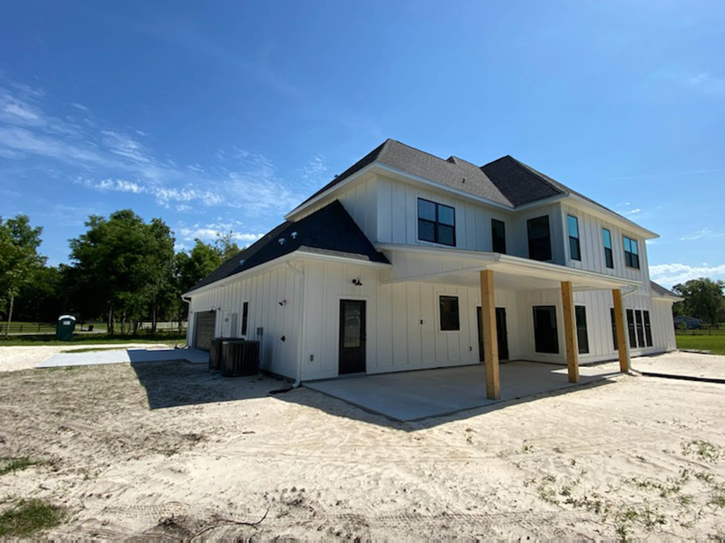 Wide front porch with white columns, dark wood door with glass window, large windows, gray siding, leafy tree in foreground, cloudy sky above