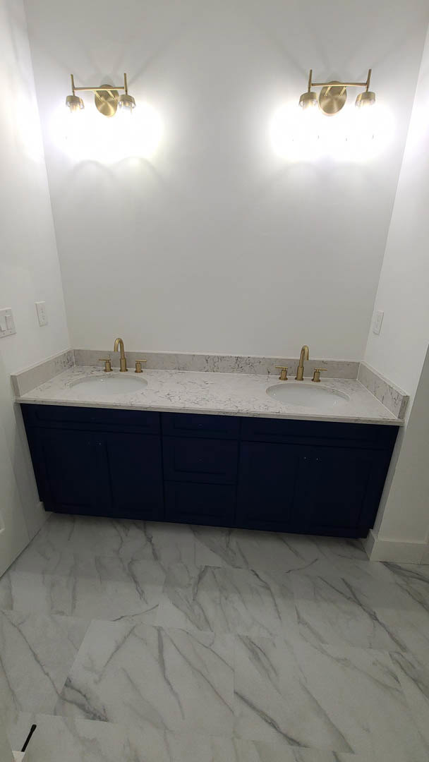 Marble countertop with dual sinks, blue vanity drawers, large wall mirror, white tile flooring, modern light fixture with wooden accent, glass shower enclosure