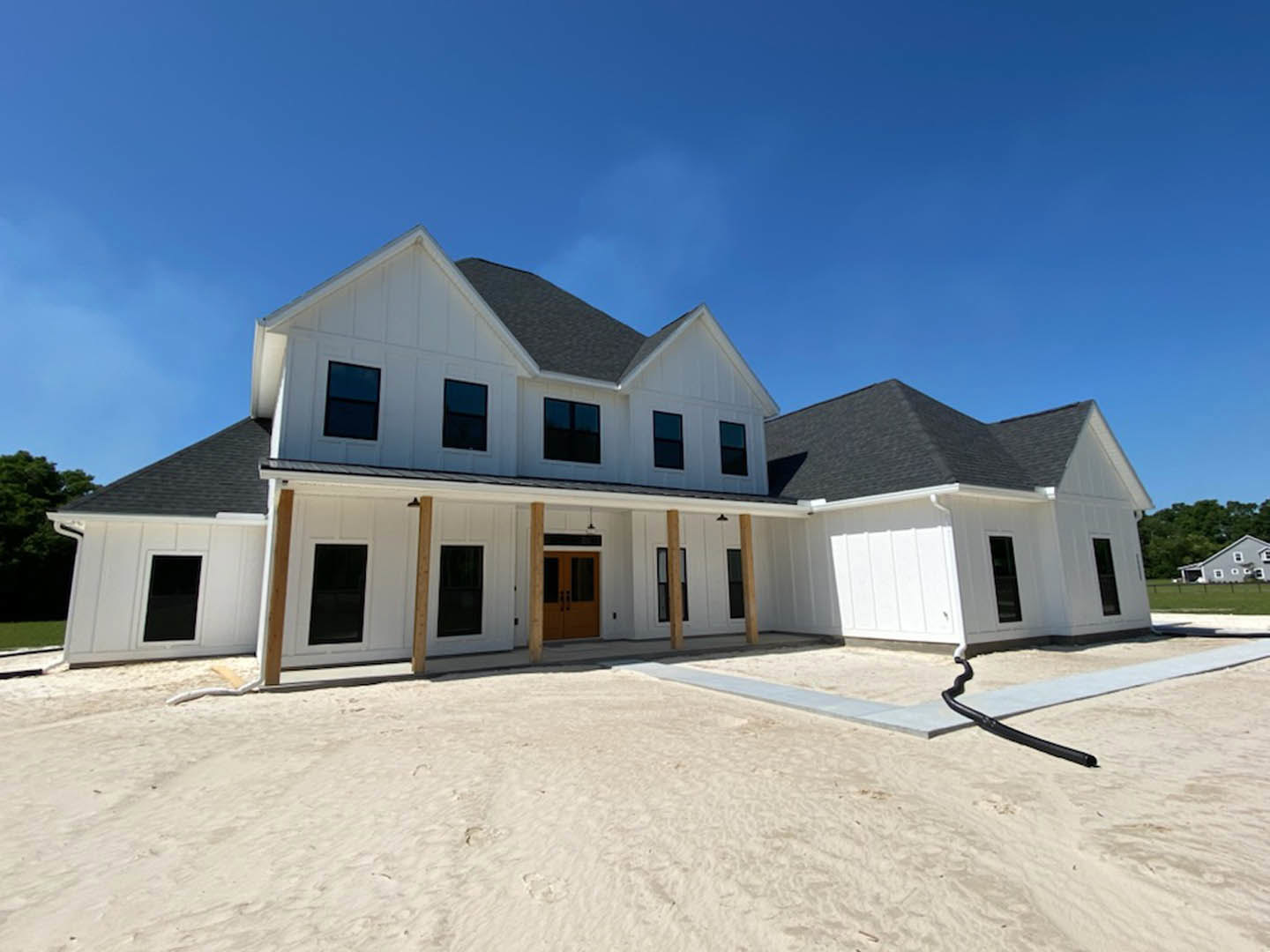 White two-story house with black roof, brown doors, and multiple windows, set against a clear blue sky; concrete surface with black pipe and sandy ground with footprints in
