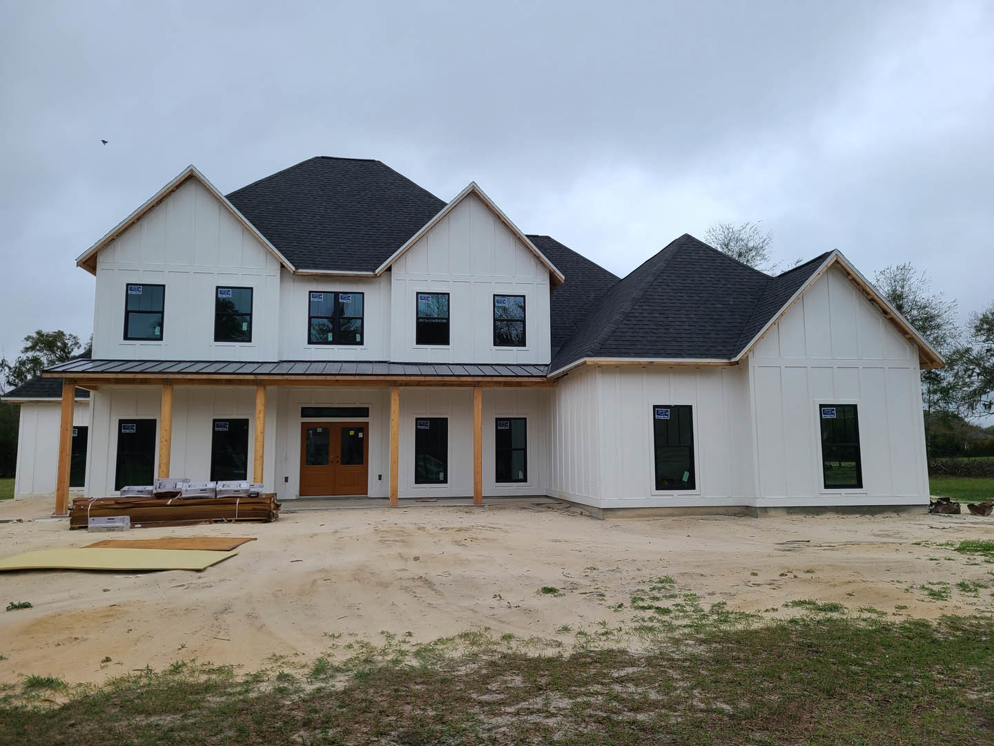White house with black roof under construction, double glass doors and windows visible, dirt patch and unpaved road in front, porch area unfinished.