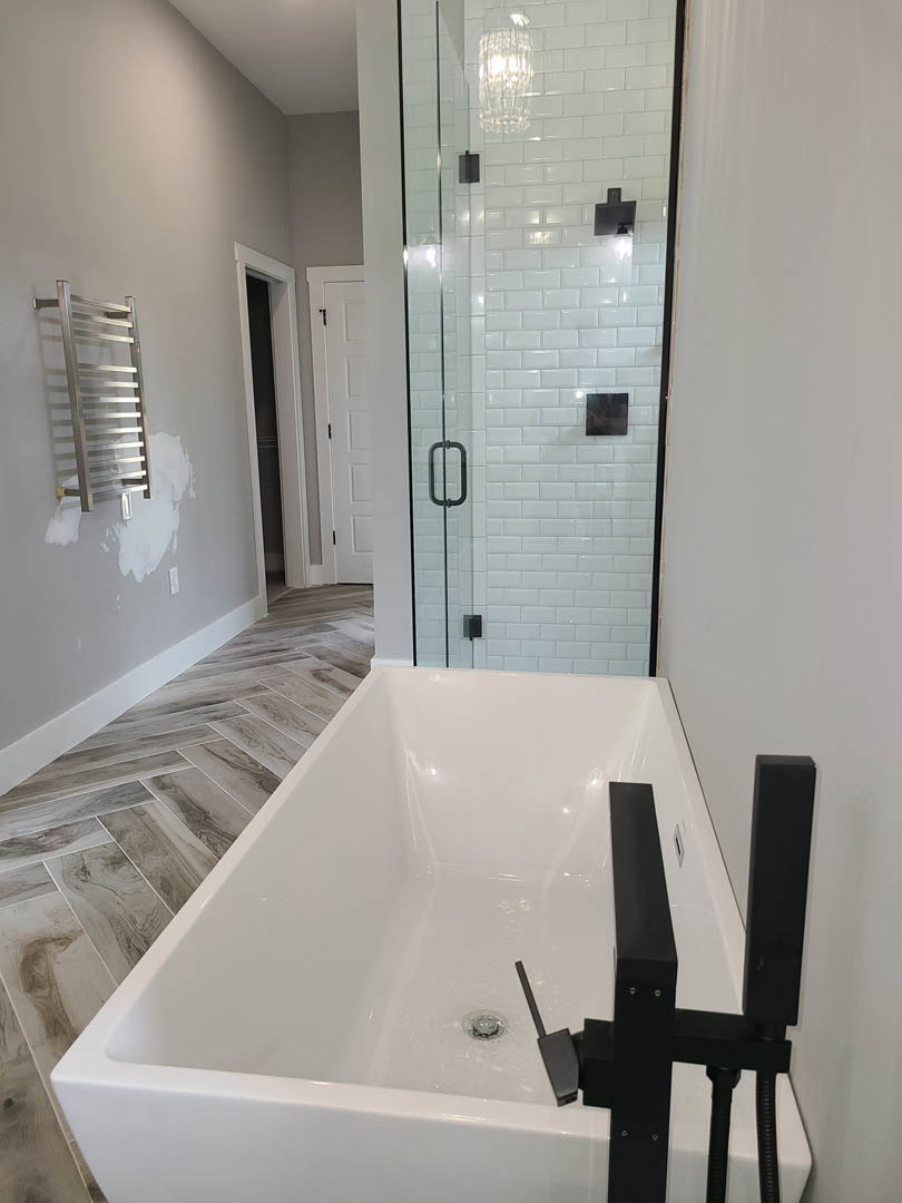 Bathroom with white bathtub featuring black handle, glass shower enclosure, wall-mounted light fixture, white door with black hinge, and tiled walls
