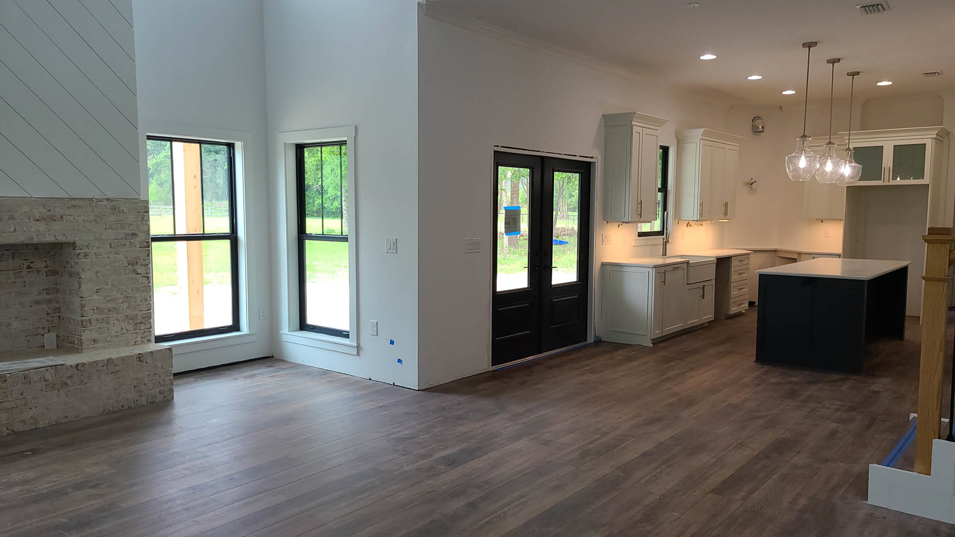 White-walled room with wood flooring, black cabinets, double glass doors, black rectangular table with white top, windows overlooking grass and trees.