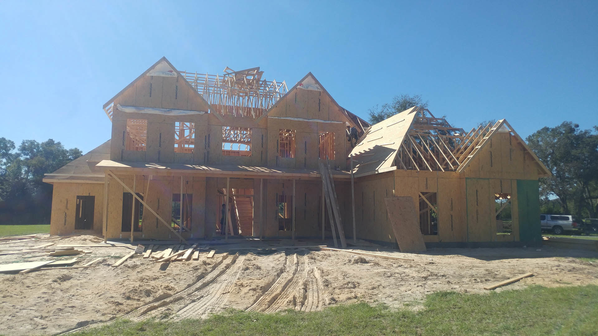 Framed house under construction with exposed roof, dirt path showing tire tracks, patches of grass, two workers on site, and a silver truck parked nearby