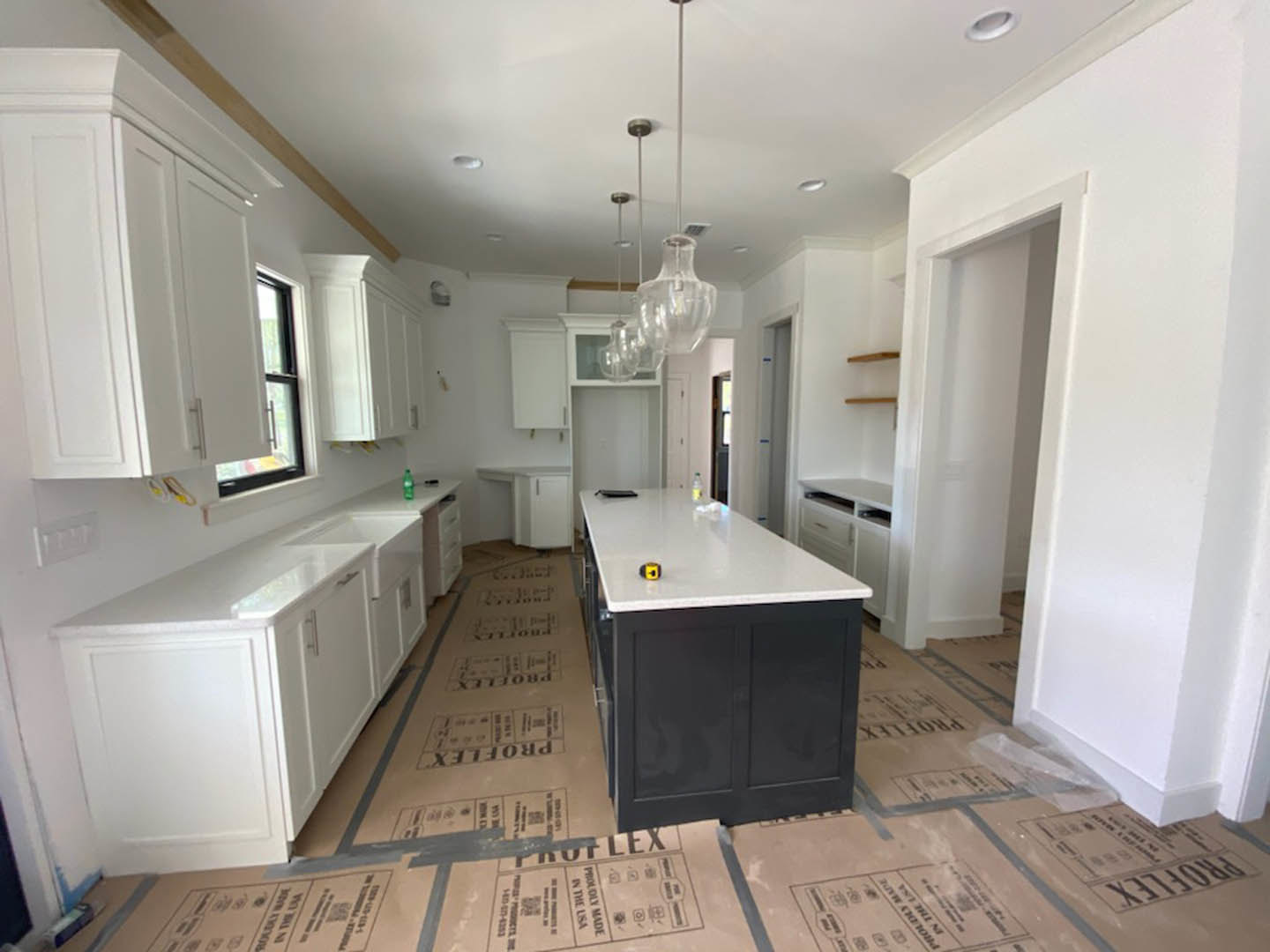 White kitchen with shaker cabinets, quartz countertop, central island with tape measure, stainless steel sink, large window, white paneled wall, cardboard box and utility knife on