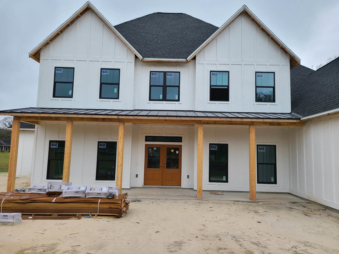 Wood-framed custom home under construction with double glass doors, black-framed windows, exposed roof beams, and stacked boxes on a pallet near the porch
