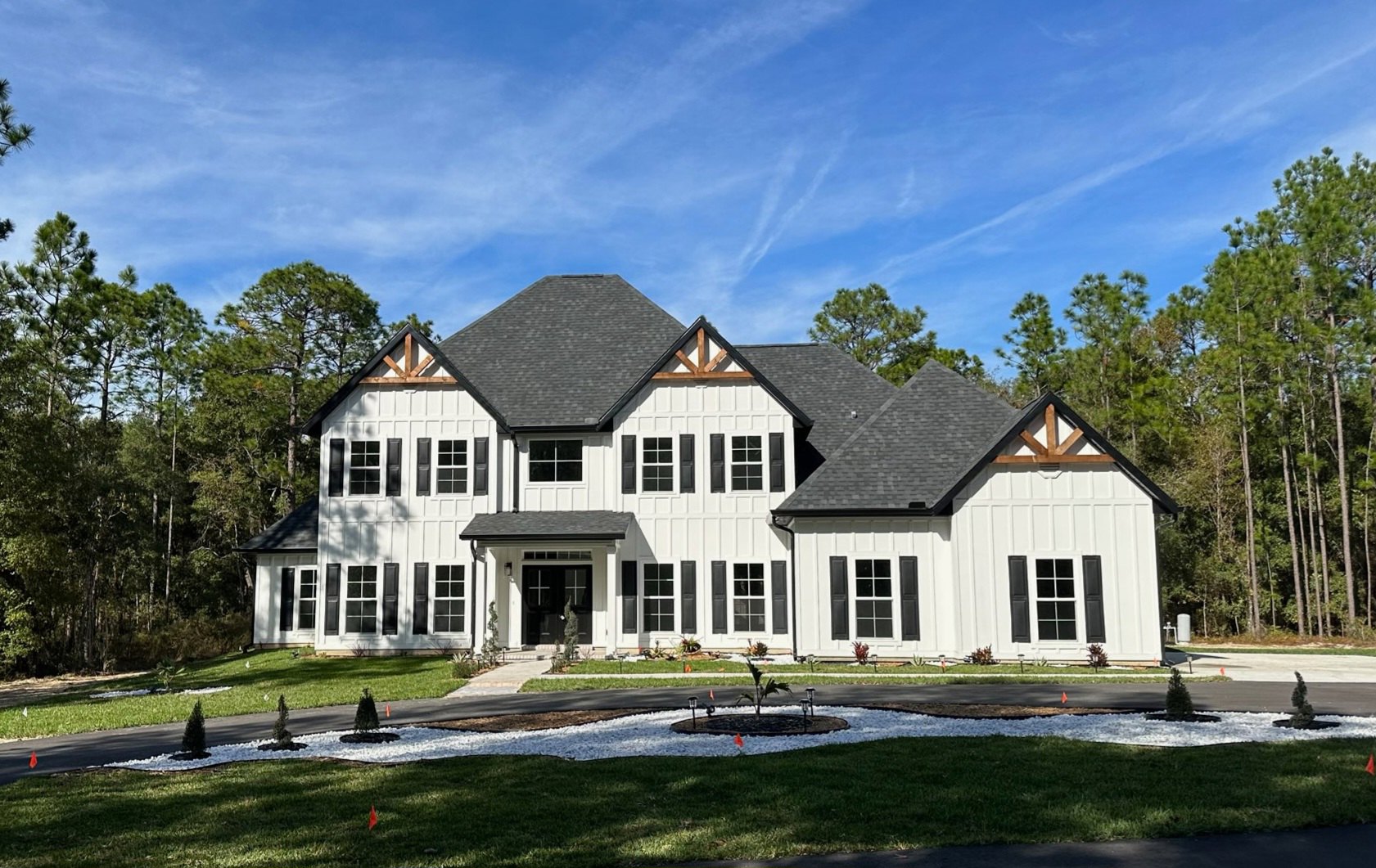 Large white house with black roof and shutters, wide driveway bordered by small white rock garden, mature trees lining the property, blue sky with scattered clouds