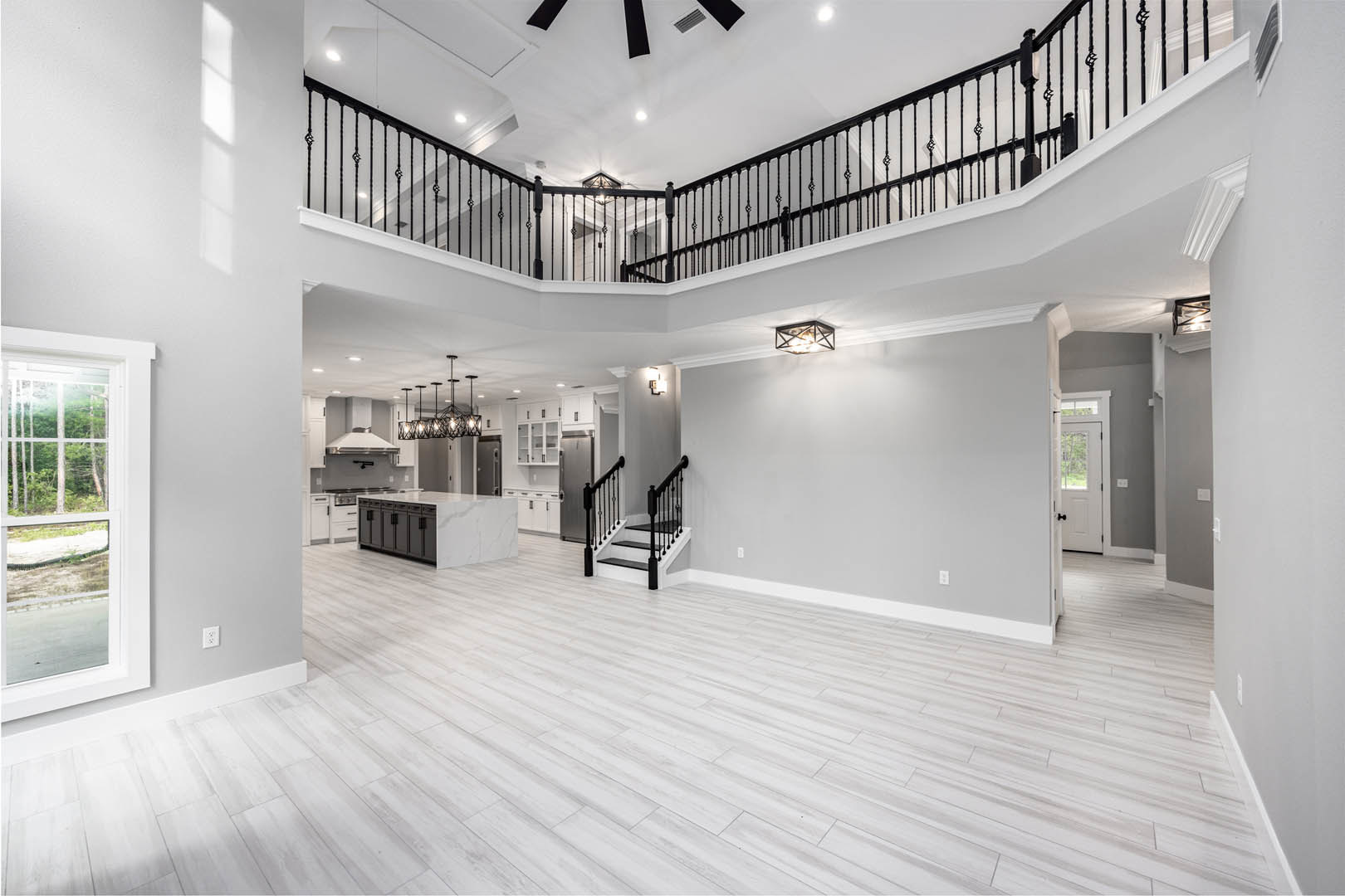 Spacious open-concept room featuring a double-story staircase with black railings, white wood flooring, modern kitchen with cabinetry, large windows overlooking trees, and