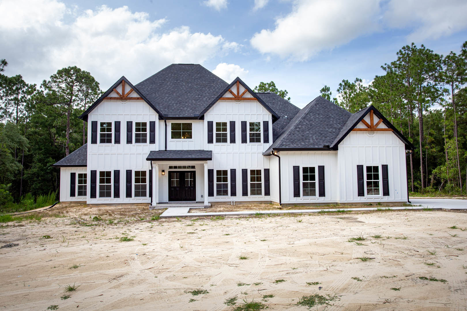 White two-story house with black shutters, black roof, and double white-framed front doors; dirt driveway with tire tracks in foreground; blue sky with scattered clouds above.