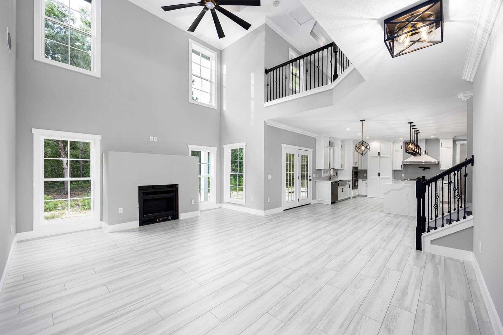Spacious white living room featuring black fireplace set in white wall, wood laminate flooring, ceiling fan with light, metal-framed light fixture, window overlooking tree, and