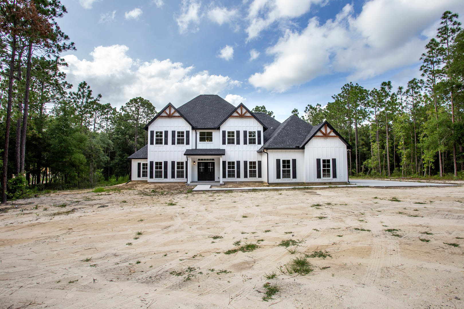 White two-story house with black roof, black double doors featuring white cross accents, large windows, dirt patch in front yard, surrounded by tall trees under a blue sky with