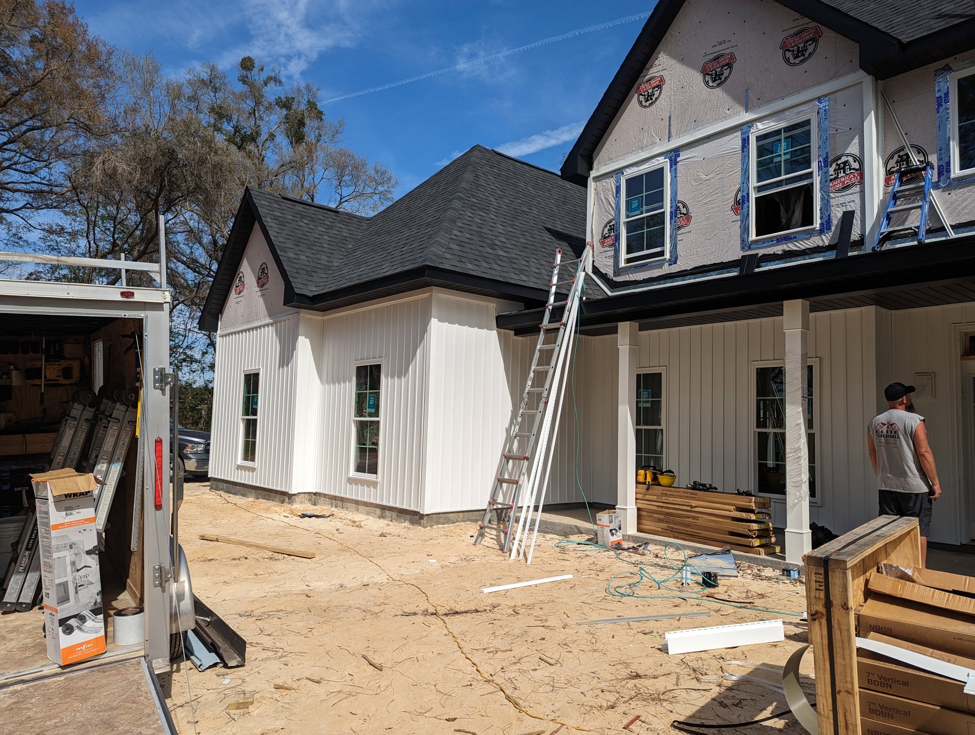 Partially built home with exposed framing, ladder leaning against exterior wall, man in grey shirt near wooden crate and cardboard boxes, construction materials scattered on