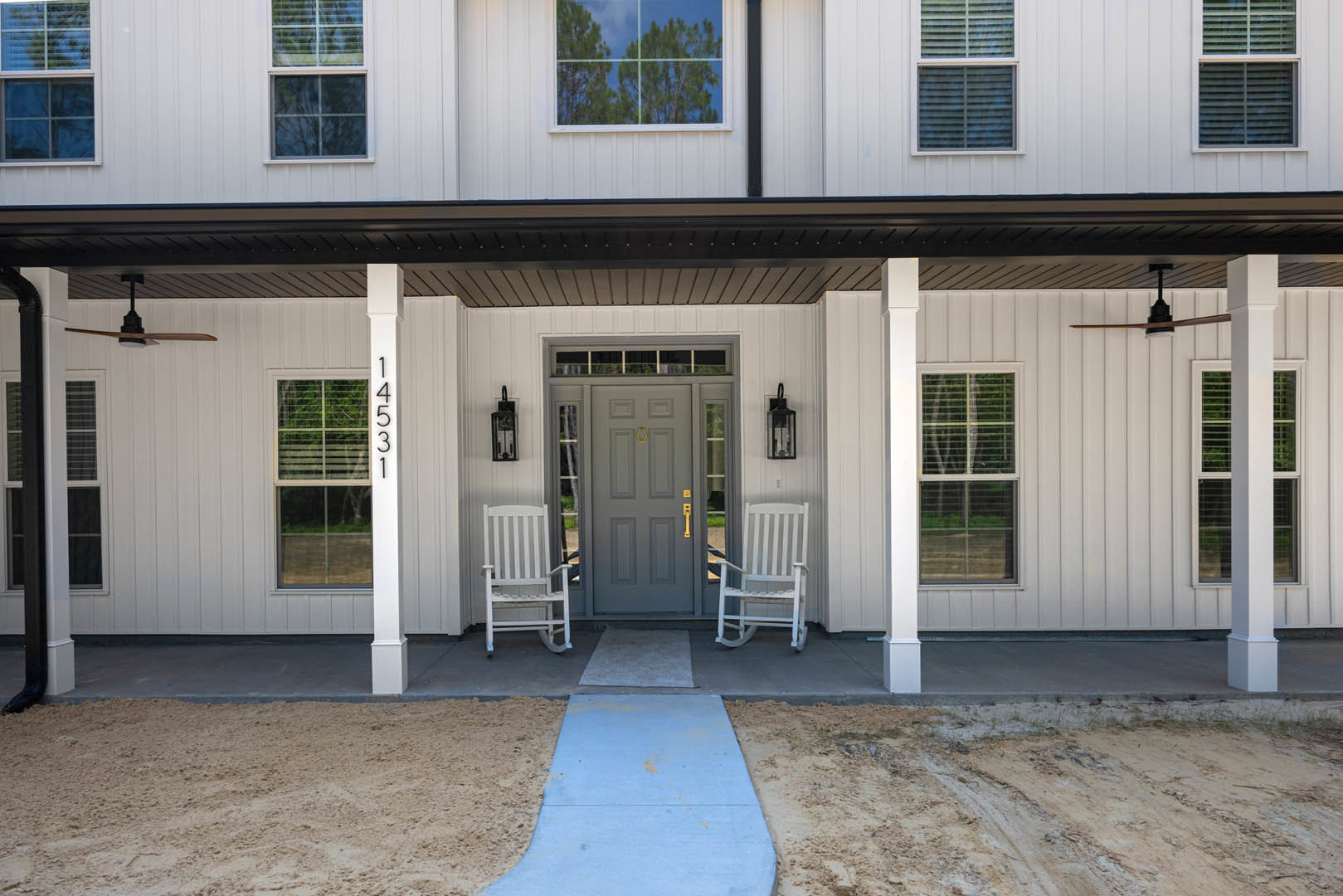 White exterior home with two white rocking chairs on a porch, grey door with glass panes, striped siding, blue concrete walkway, and sandy ground.