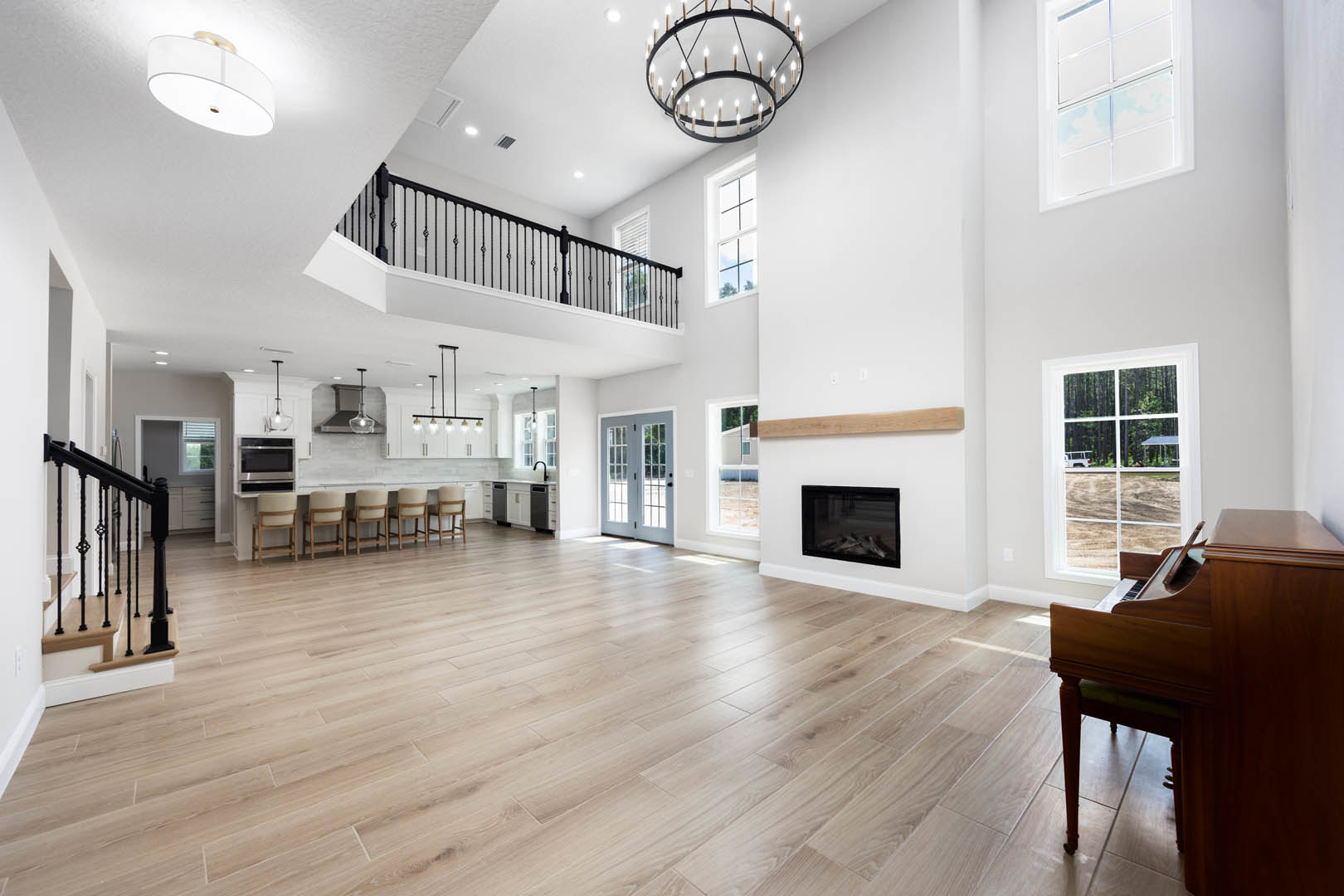 Spacious white interior with hardwood floors, chandelier, staircase, glass-framed black accent, piano, and white chair with wooden frame