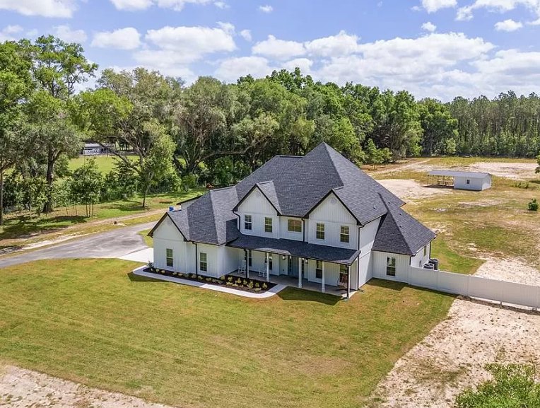 Large white house with black roof, expansive green lawn, mature trees in background, blue sky with scattered clouds, dirt road bordered by grass and wooden fence