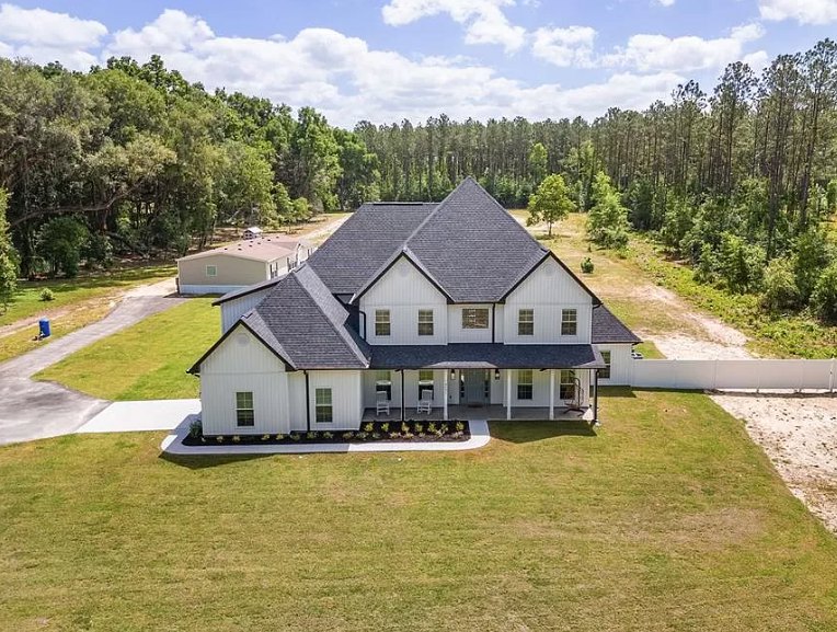 White two-story house with black roof, multiple windows, manicured green lawn, mature trees in background, blue sky with scattered clouds