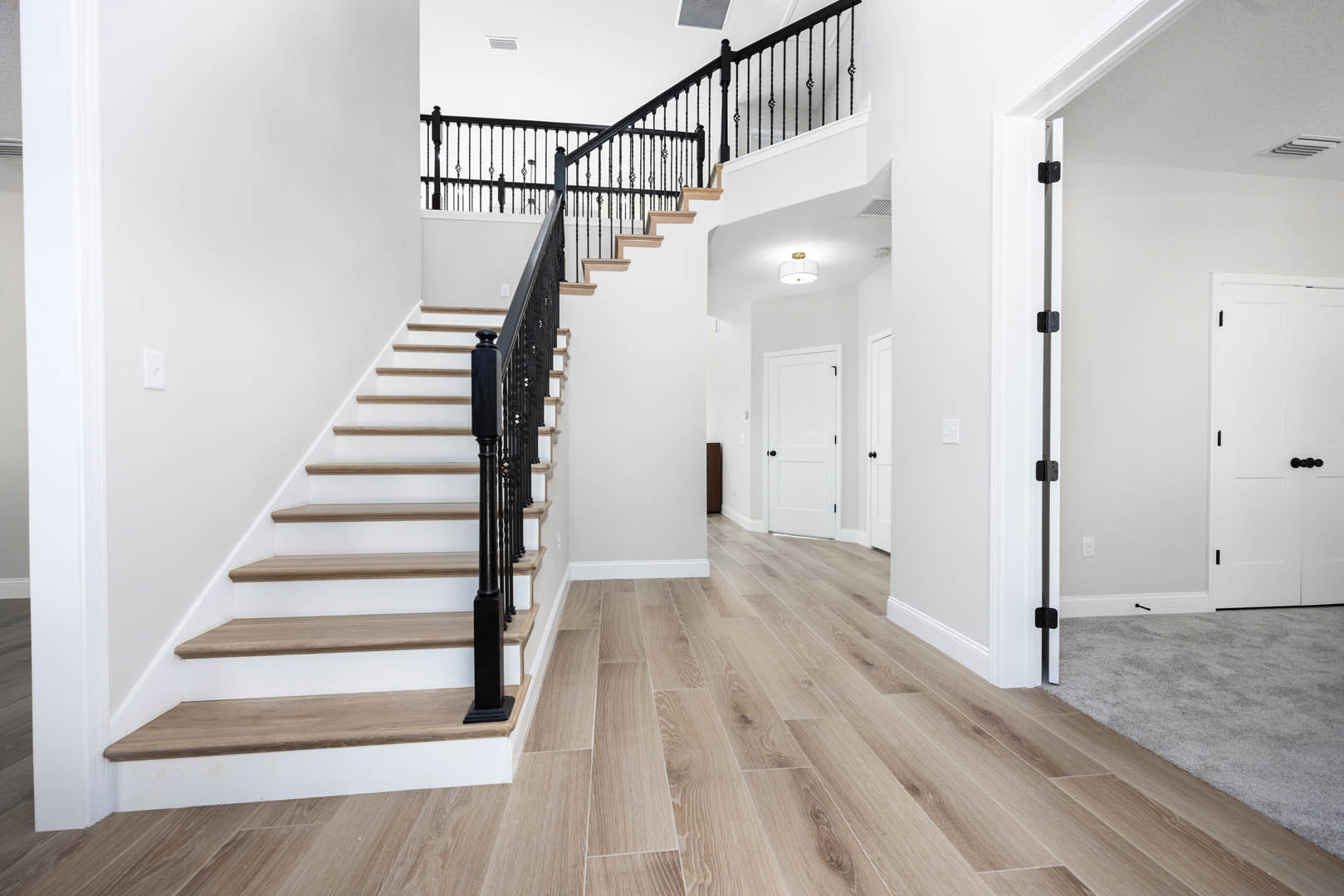 White staircase with natural wood steps and matching handrail, white paneled door with black hardware, beige carpet runner, hardwood flooring in hallway, smooth white walls and