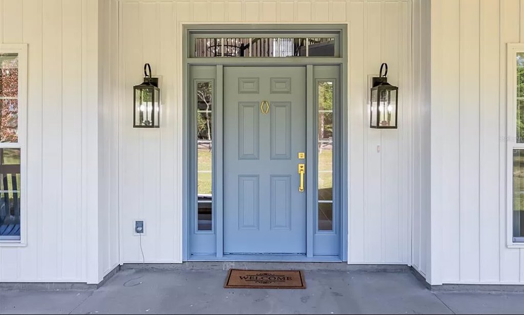 Blue front door with gold handle, flanked by black metal sconces, and a textured doormat on concrete porch.