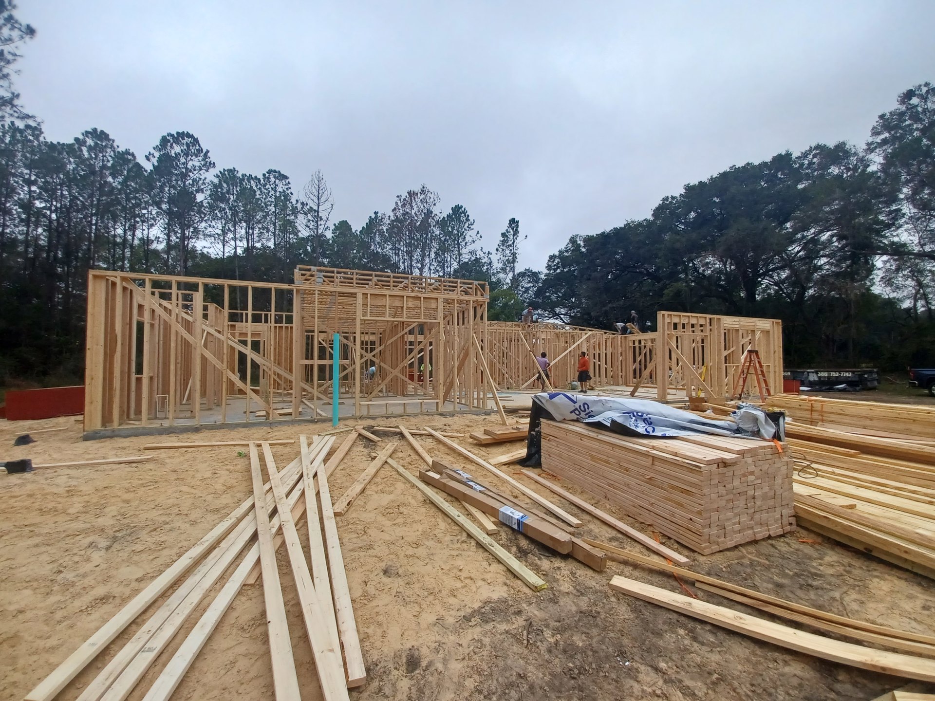 Framed wooden beams and stacked planks at a residential construction site, foundation visible with trees in the background