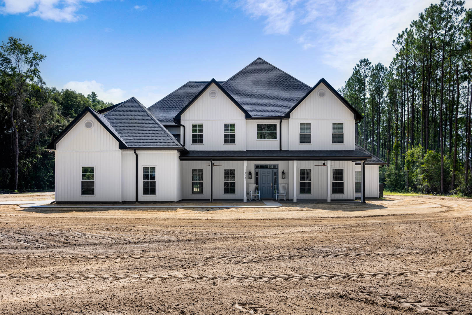 White two-story house with blue front door, covered porch, white-framed windows, and gabled roof; dirt road with tire tracks in foreground; trees and cloudy sky in background.
