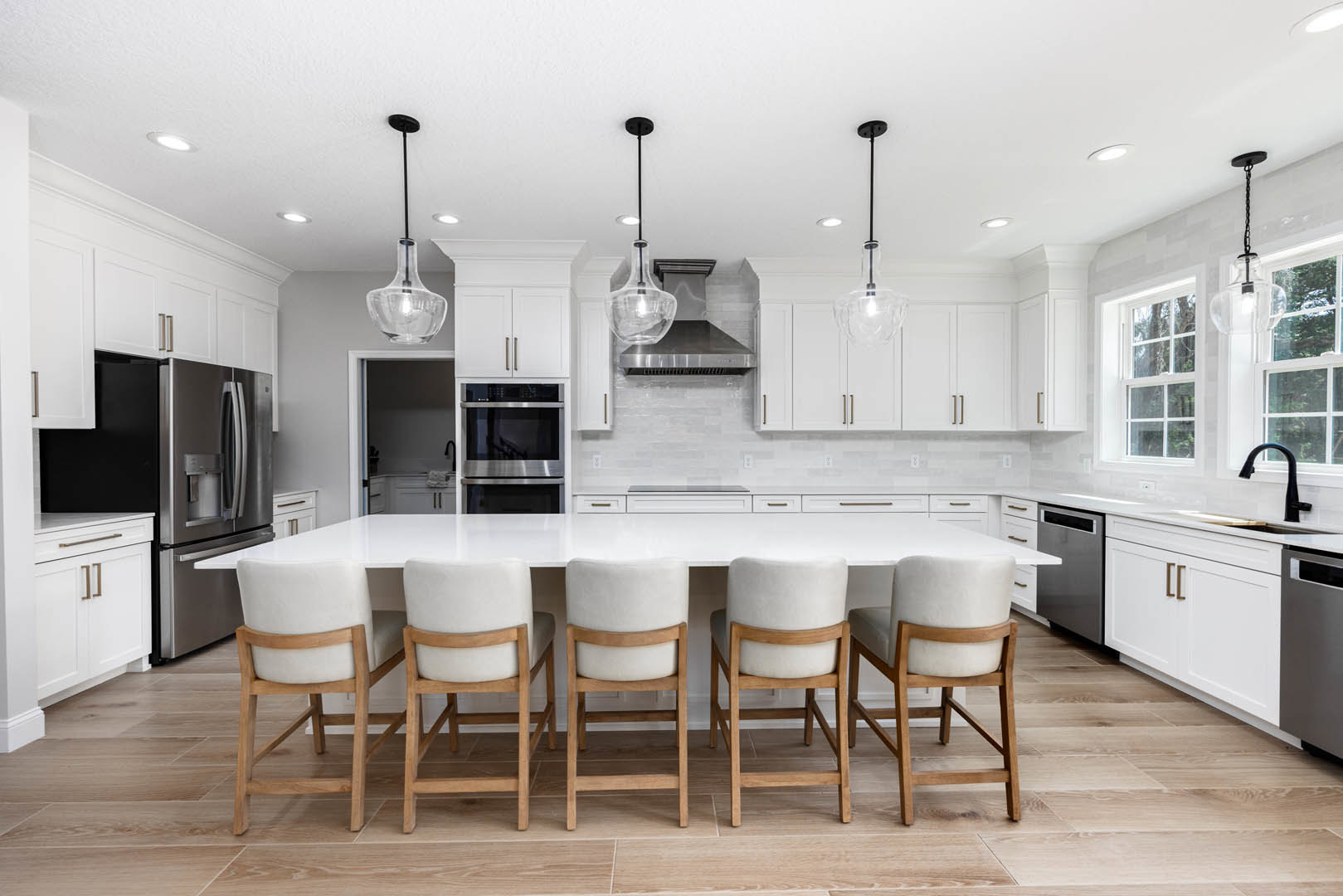 Spacious kitchen featuring a wooden dining table surrounded by white chairs with wooden legs, double wall ovens, light fixtures hanging from the ceiling, glass containers on the