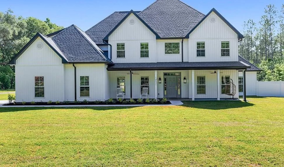 Large white house with grey door and glass panes, expansive green lawn, black pole near porch, Robert Frost Farm visible in background, trees and blue sky overhead.