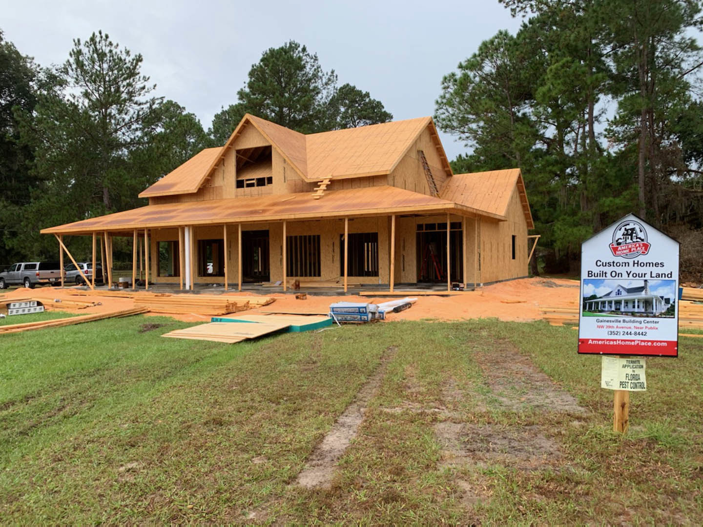 Wood-framed house under construction surrounded by tall trees, unfinished porch, silver pickup truck parked on grassy lot, white sign with black text near scattered lumber