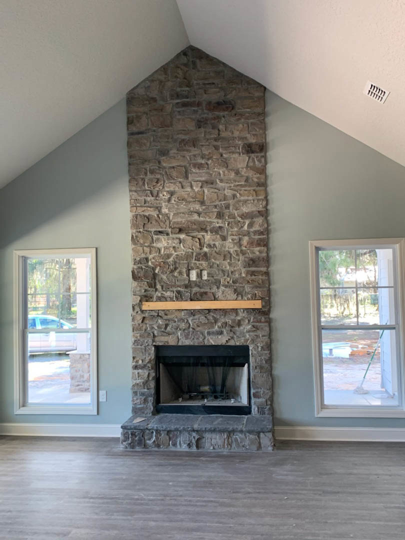 Stone fireplace with black and white surround, wood shelf mantel, concrete floor, large window showing car and ladder outside