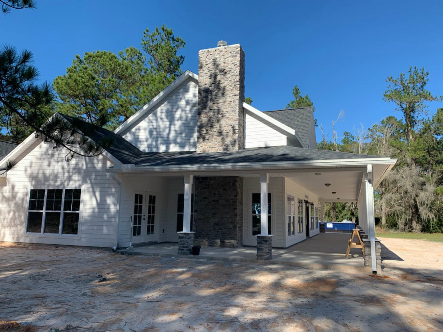 White siding house with prominent stone chimney, stone pillar, and window; wooden ladder rests on porch near stone wall; trees and sky in background.