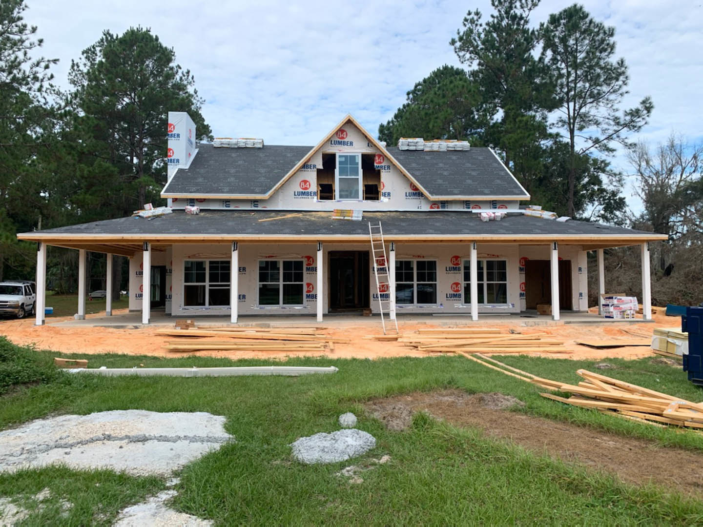 Wood-framed house under construction with triangular roof, ladder leaning against wall, white car parked nearby, white-framed window, grassy yard, Karen Blixen Museum visible in