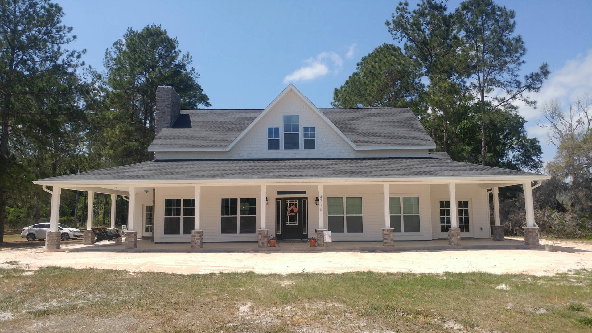 Spacious front porch with white columns, black door adorned with a wreath, white-framed windows, grassy yard, and a smaller white house visible in the background.